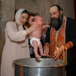 A baby being baptized by a priest and woman in a traditional church setting.